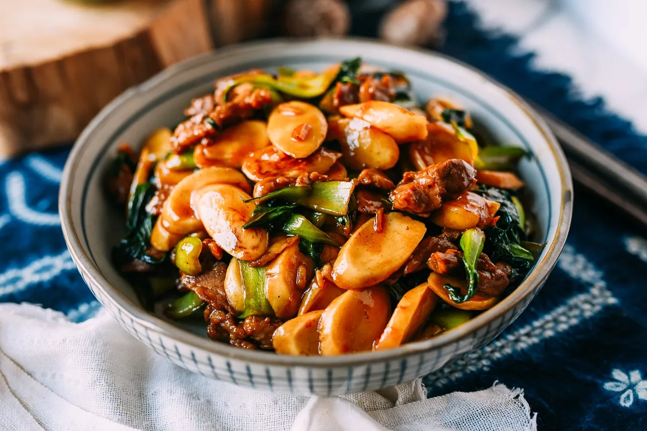Stir-fried rice cakes with vegetables and meat in rich sauce, served in bowl on blue-white patterned cloth
