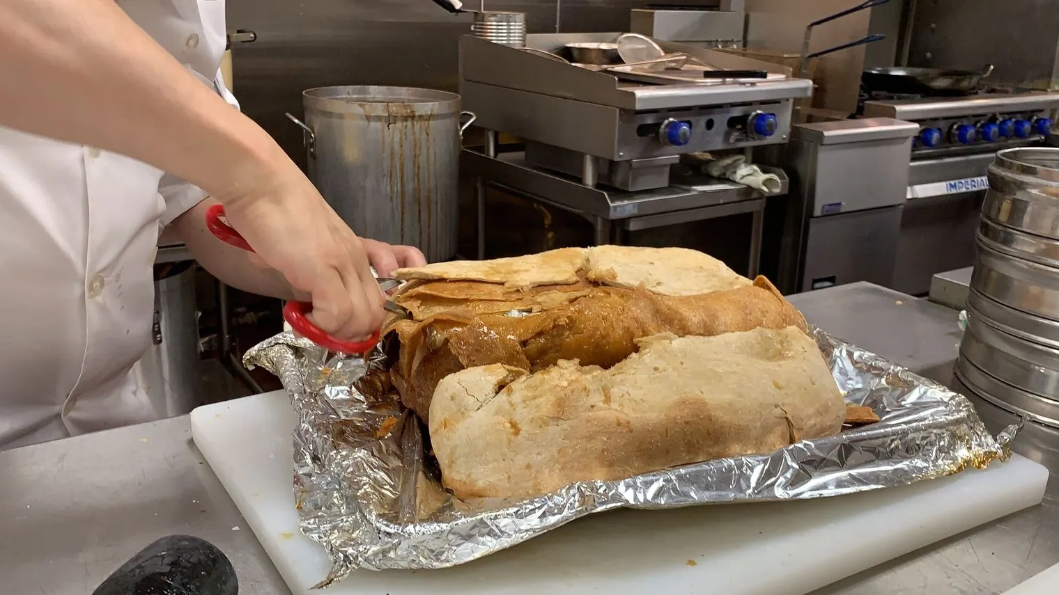 Chef in white coat cutting beggars chicken with red scissors on foil-lined board