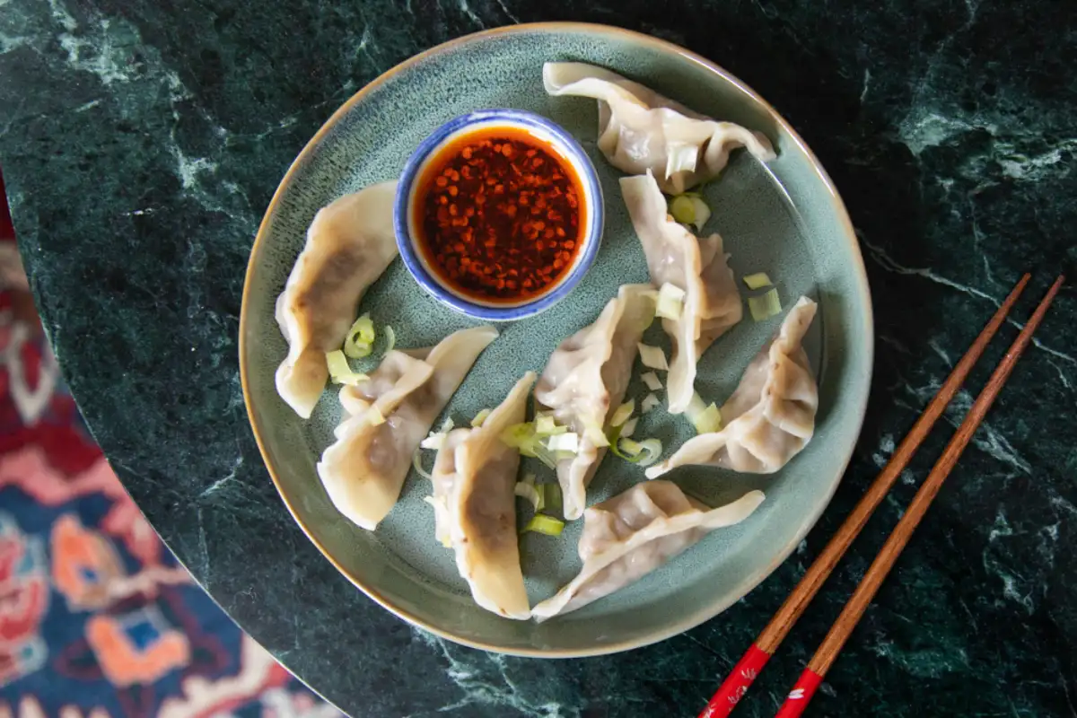 Traditional Chinese dumplings with chili dipping sauce, garnished with scallions, served on dark marble surface with red chopsticks