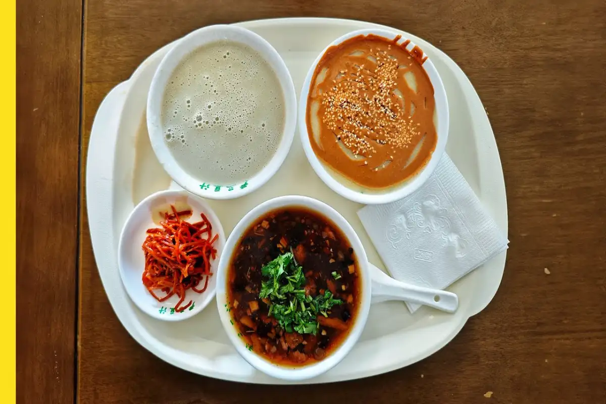 Breakfast tray with four dishes: douzhi, sesame-topped brown paste, shredded pickled carrots, and herbed dark broth on wooden table