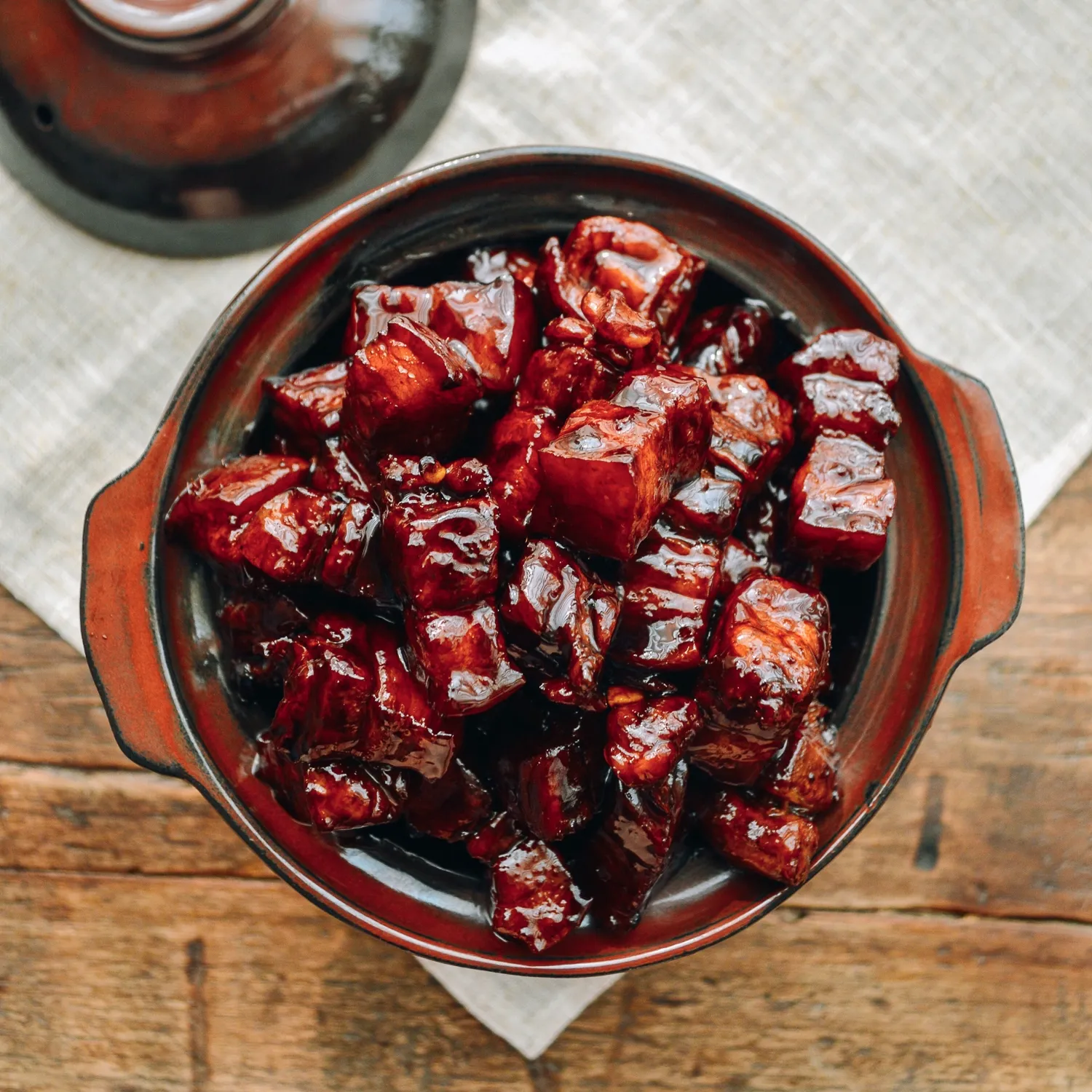Tender braised pork cubes in glossy sauce, served in a bowl on wooden table with lid in background